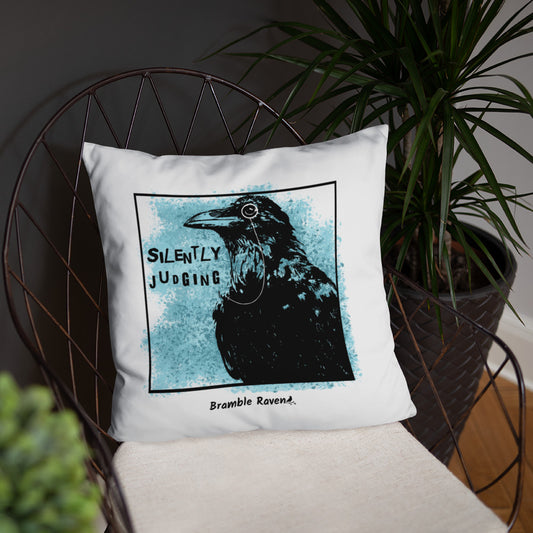18 by 18 inch accent pillow with silently judging text by black crow wearing a monocle in a square with blue paint splatters on a white background.  Design on both sides of pillow. Shown on wicker chair.