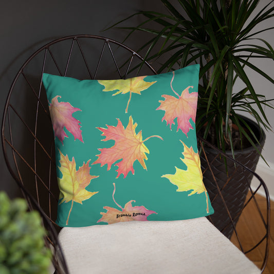 18 by 18 inch accent pillow featuring watercolor fall leaves on a dark green background. Pillow is double-sided. Larger patterned leaves on the back. Smaller patterned leaves on the front. Shown on a wicker chair.