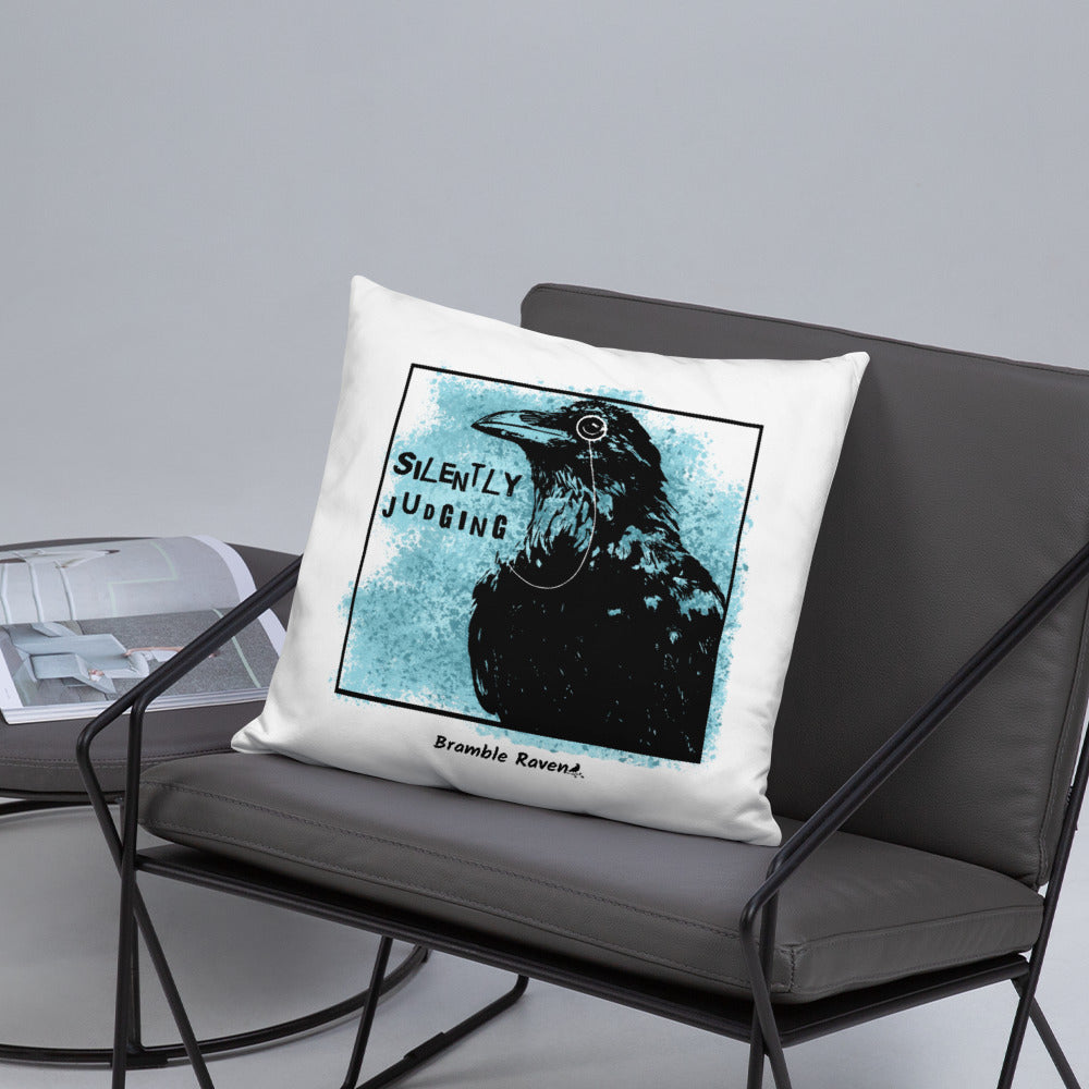 18 by 18 inch accent pillow with silently judging text by black crow wearing a monocle in a square with blue paint splatters on a white background.  Design on reverse side of pillow doesn't have paint splatters. Shown on grey chair.