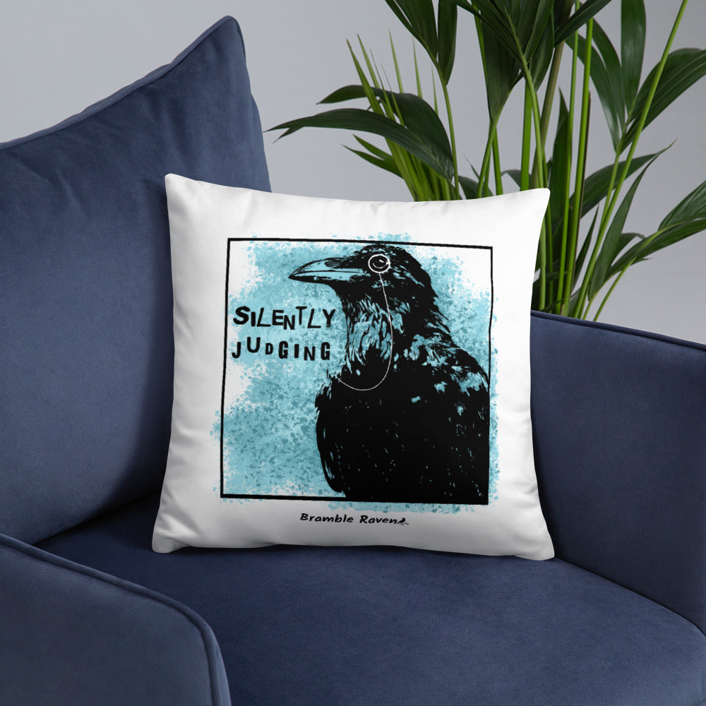 18 by 18 inch accent pillow with silently judging text by black crow wearing a monocle in a square with blue paint splatters on a white background.  Design on reverse side of pillow has no splatters. Shown on blue sofa.