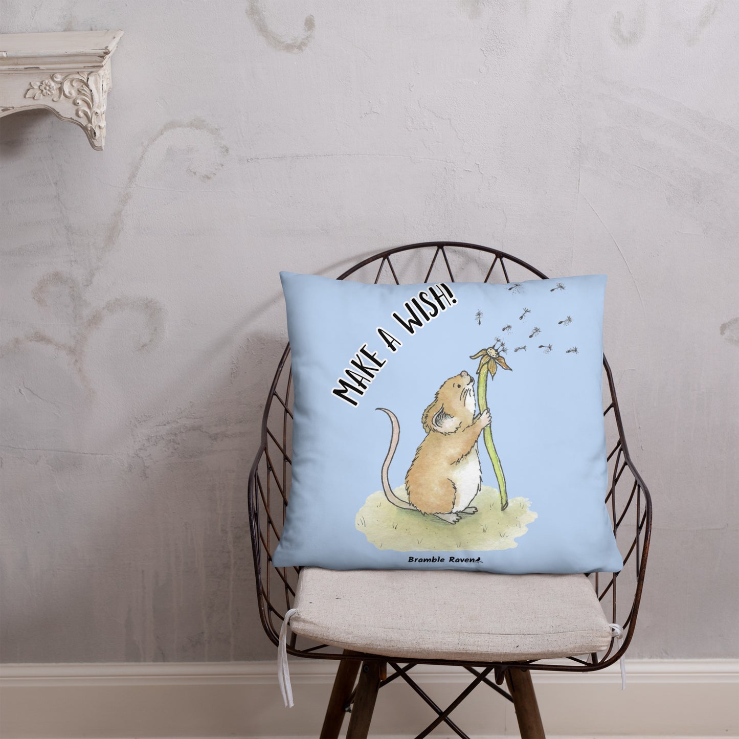 Original Dandelion Wish design of cute watercolor mouse blowing dandelion seeds on a light blue background. Double-sided image on 22 by 22 inch pillow. Back image has make a wish phrase. Shown on wicker chair.