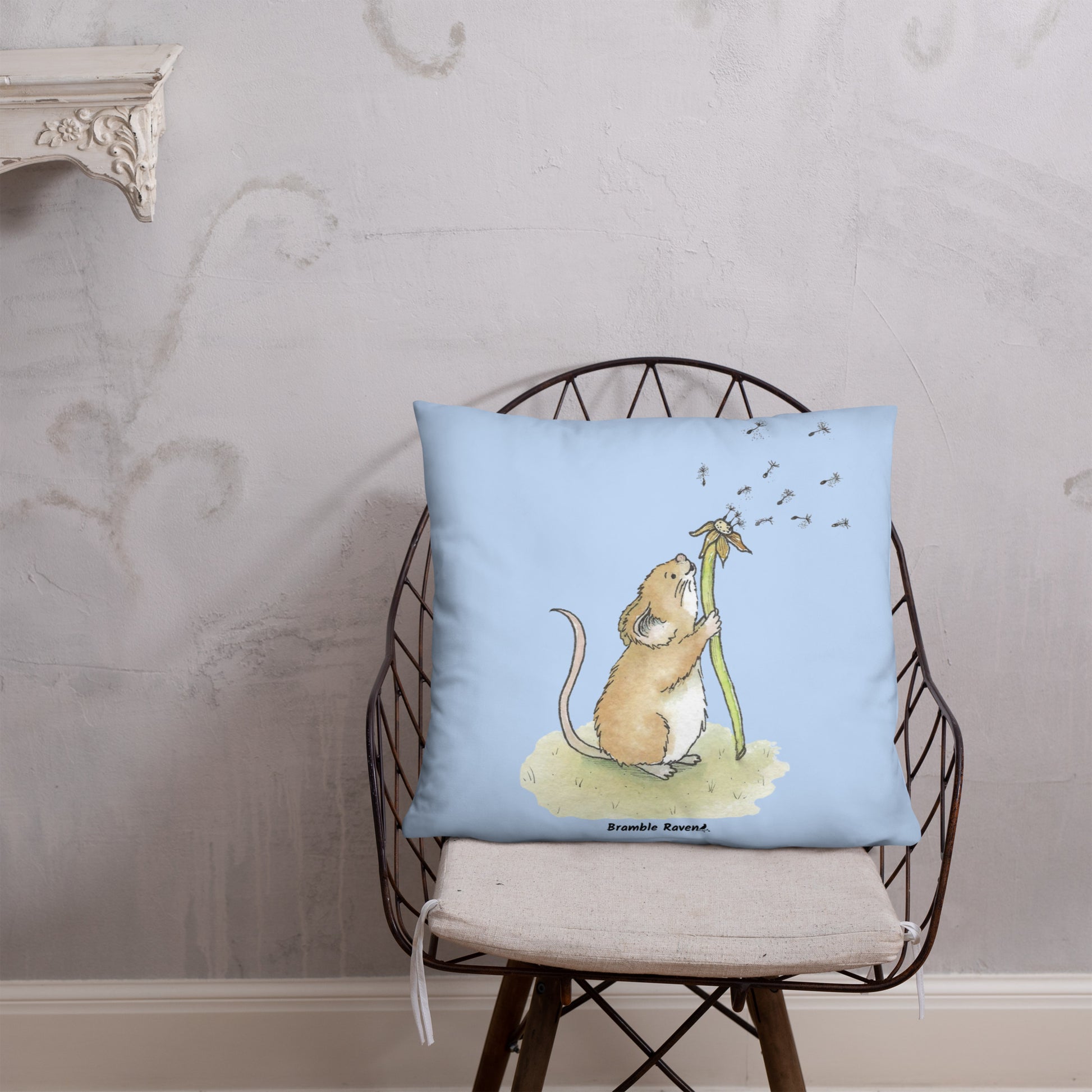 Original Dandelion Wish design of cute watercolor mouse blowing dandelion seeds on a light blue background. Double-sided image on 22 by 22 inch pillow. Shown on wicker chair.