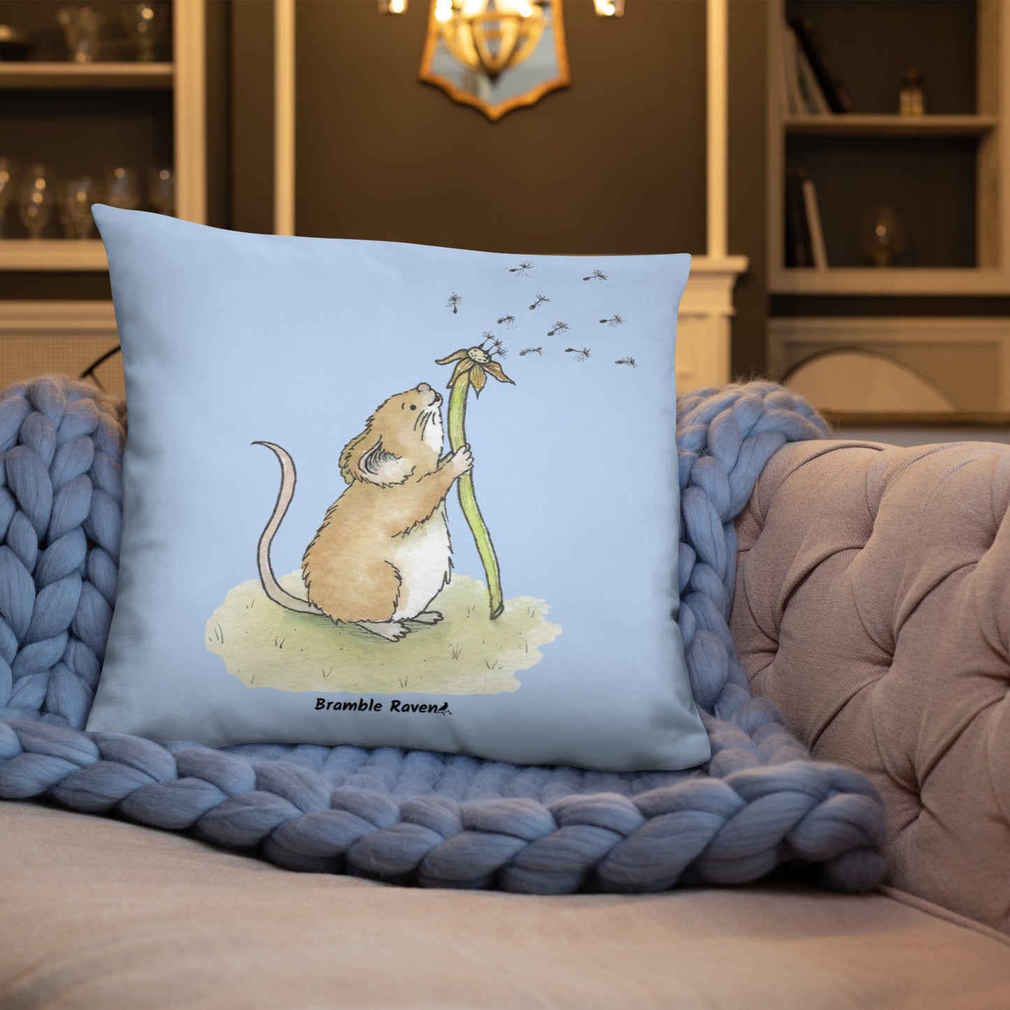 Original Dandelion Wish design of cute watercolor mouse blowing dandelion seeds on a light blue background. Double-sided image on 22 by 22 inch pillow. Shown on blue knitted blanket on couch.