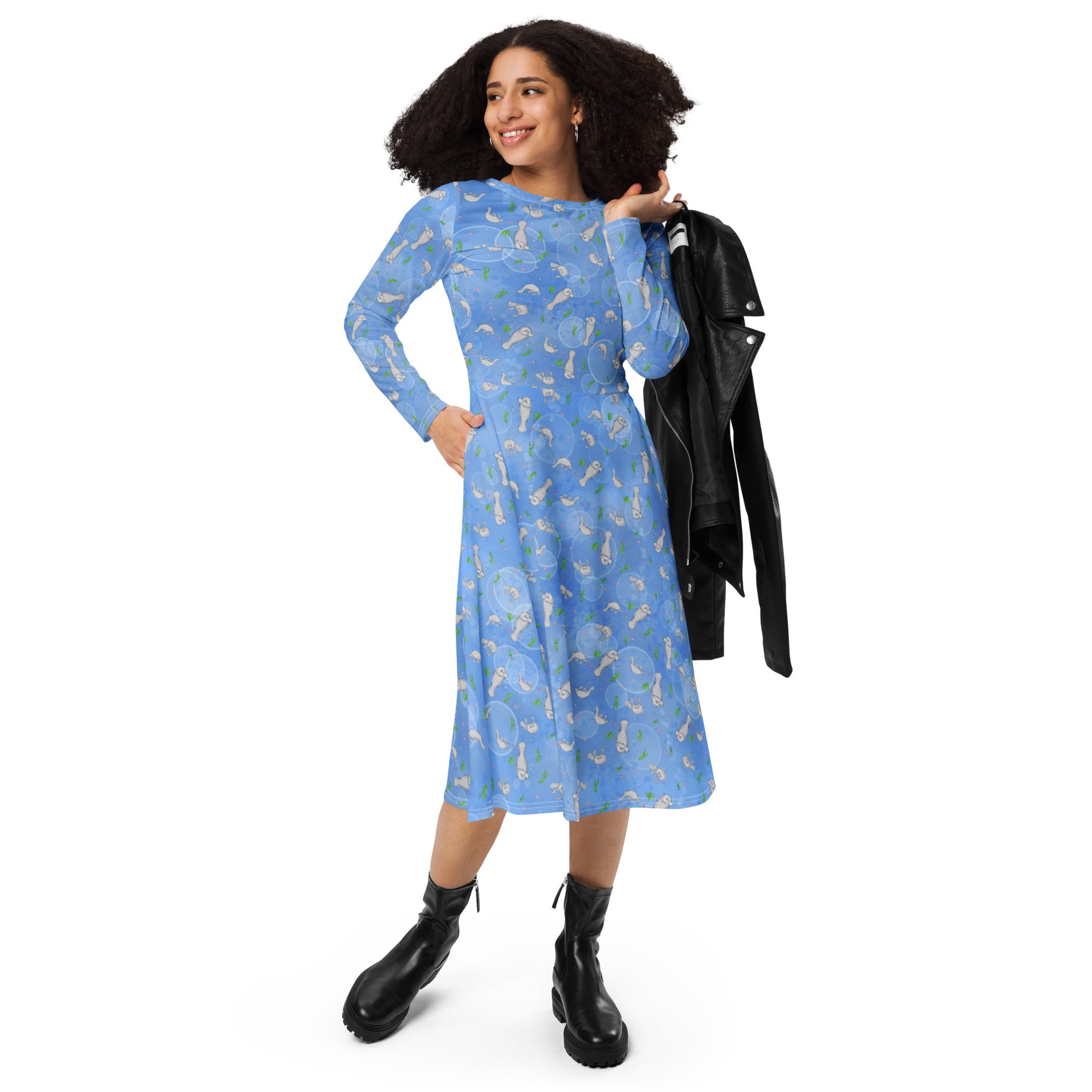 Long sleeve midi dress with fitted waist, flared bottom, and side pockets. Features a patterned design of manatees, seashells, seaweed, and bubbles on an ocean blue background. Shown on female model with black jacket slung over her shoulder.