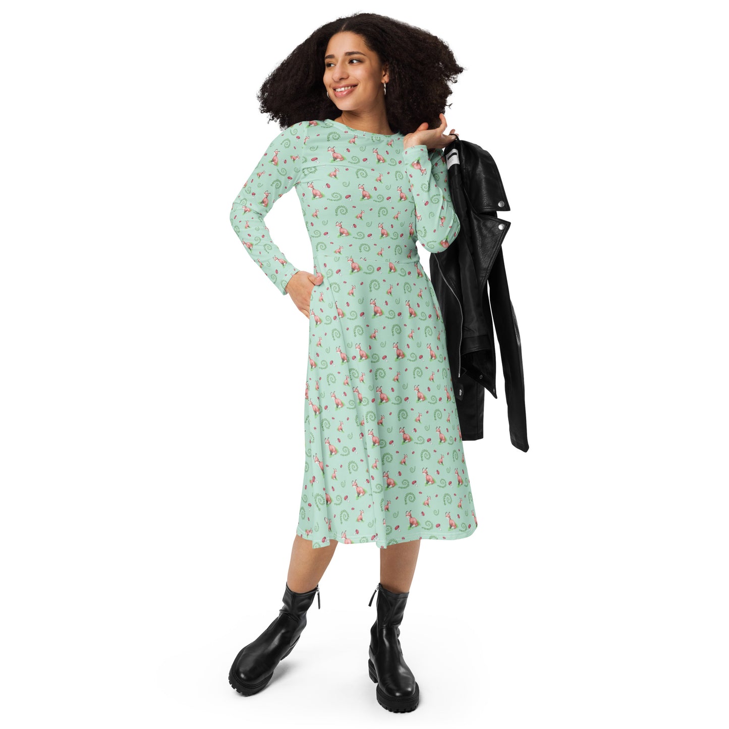 Forest fox patterned long sleeve midi dress with side pockets. Has watercolor foxes, mushrooms, and ferns on a light green fabric. Features boat neckline and fitted elastic waist. Shown on female model with black jacket on her shoulder.