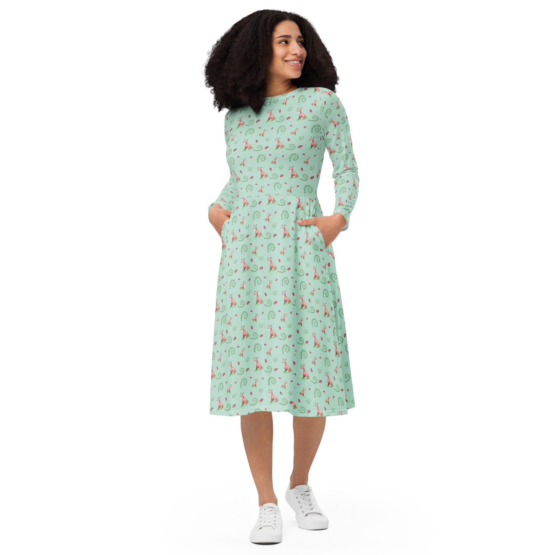 Forest fox patterned long sleeve midi dress with side pockets. Has watercolor foxes, mushrooms, and ferns on a light green fabric. Features boat neckline and fitted elastic waist. Shown on female model.