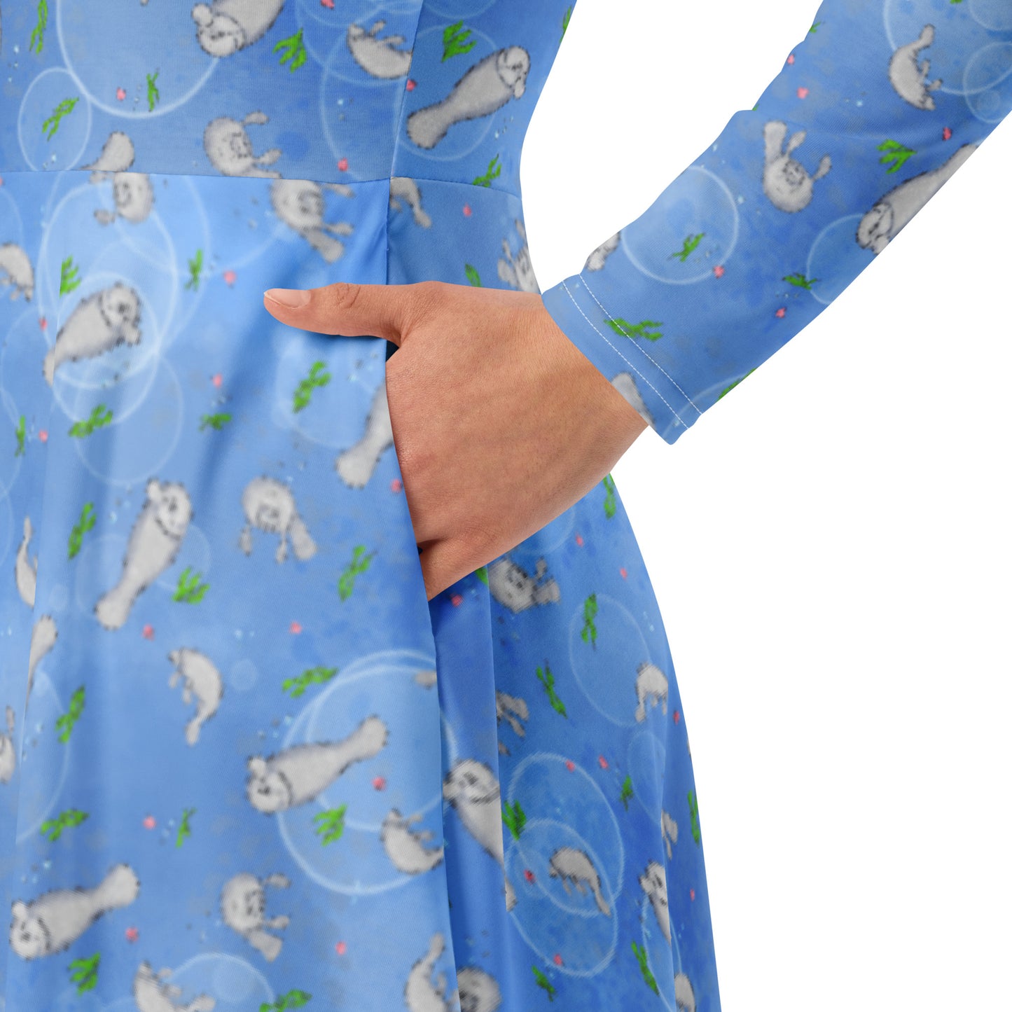 Long sleeve midi dress with fitted waist, flared bottom, and side pockets. Features a patterned design of manatees, seashells, seaweed, and bubbles on an ocean blue background. Detail image shows model's hand in pocket.