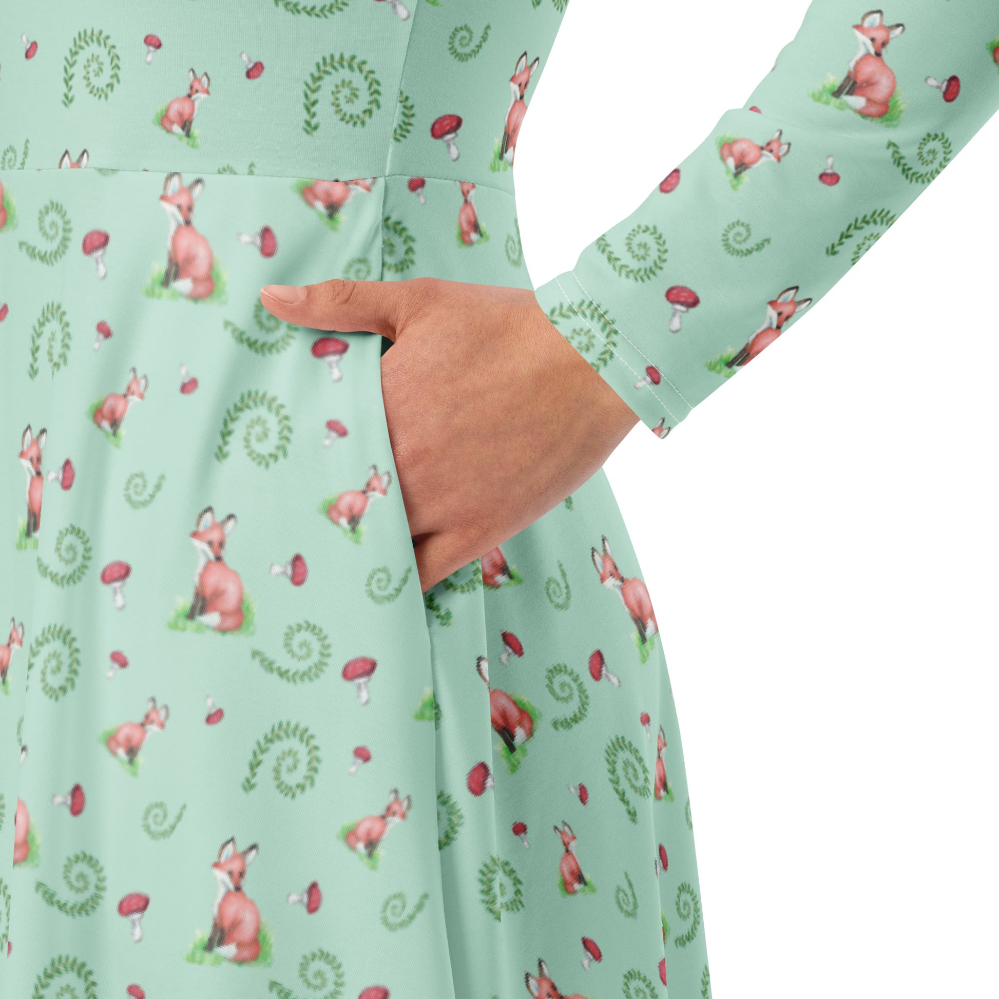 Forest fox patterned long sleeve midi dress with side pockets. Has watercolor foxes, mushrooms, and ferns on a light green fabric. Features boat neckline and fitted elastic waist. View shows close-up of hand in pocket.