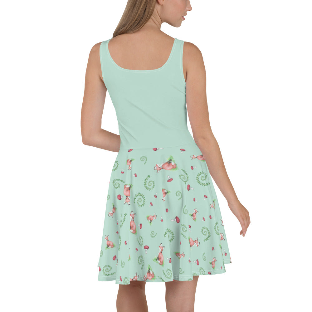 Sleeveless skater dress featuring watercolor foxes, mushrooms, and ferns on a light green fabric. Mid-thigh length flared skirt with a fitted elastic waist. Back view on female model.