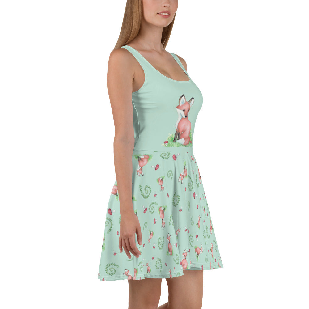 Sleeveless skater dress featuring watercolor foxes, mushrooms, and ferns on a light green fabric. Mid-thigh length flared skirt with a fitted elastic waist. Front view on female model facing right.