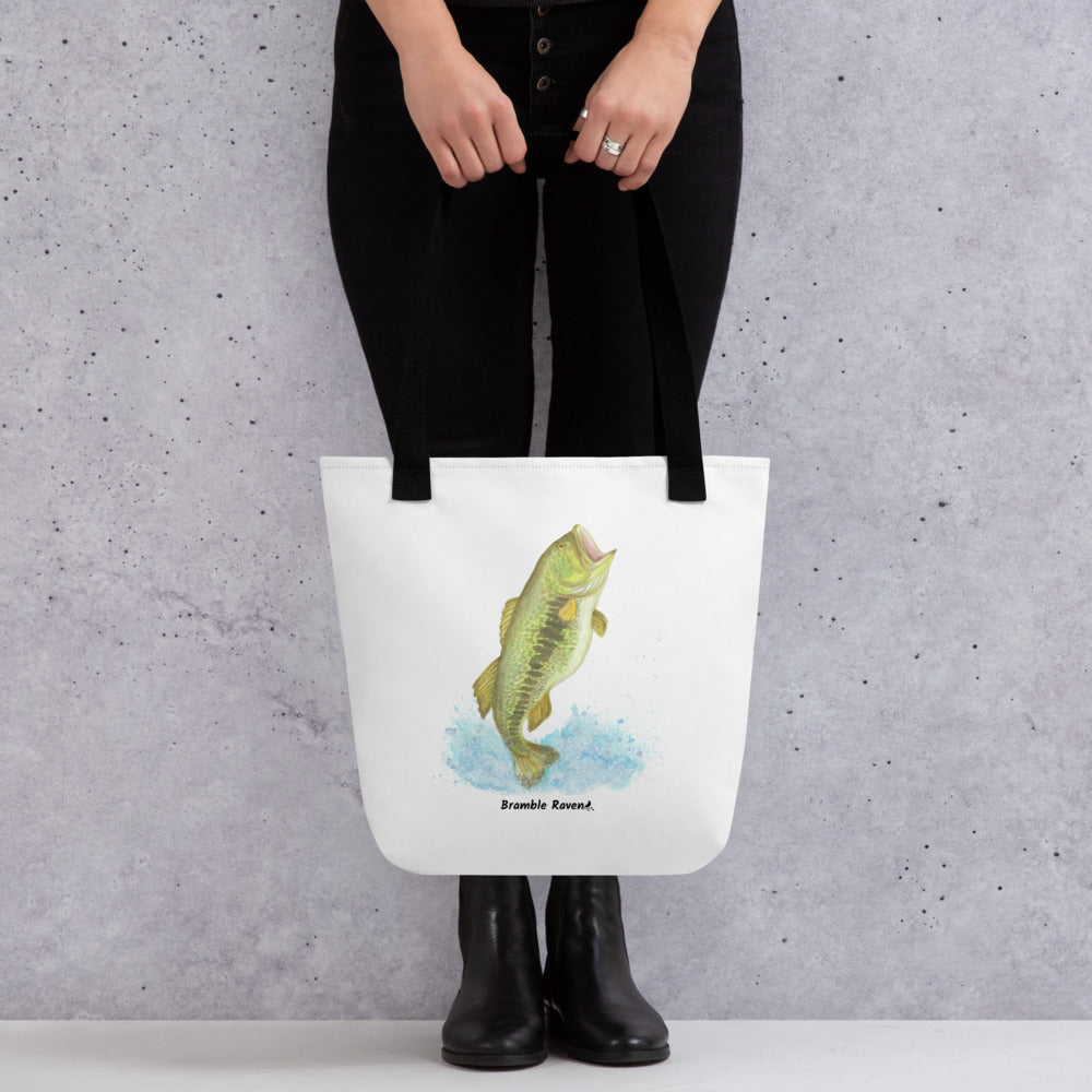 White polyester 15 by 15 inch tote bag. Features a watercolor painting of a largemouth bass leaping out of the water. Black handles. Shown hanging from a model's hands.