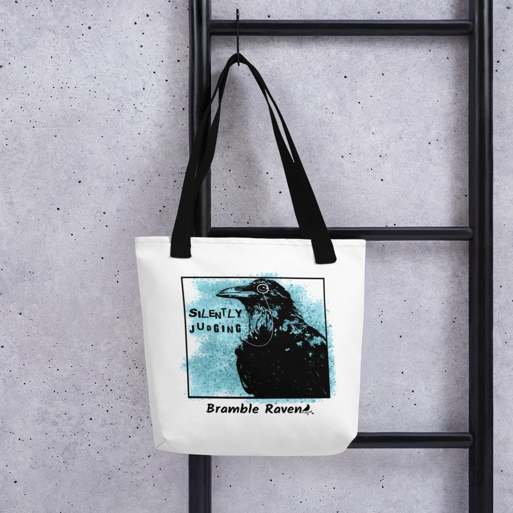 15 by 15 inch tote bag with silently judging text by black crow wearing a monocle in a square with blue paint splatters.  Design on both sides of bag. White polyester fabric with black handles. Shown hanging on black ladder.