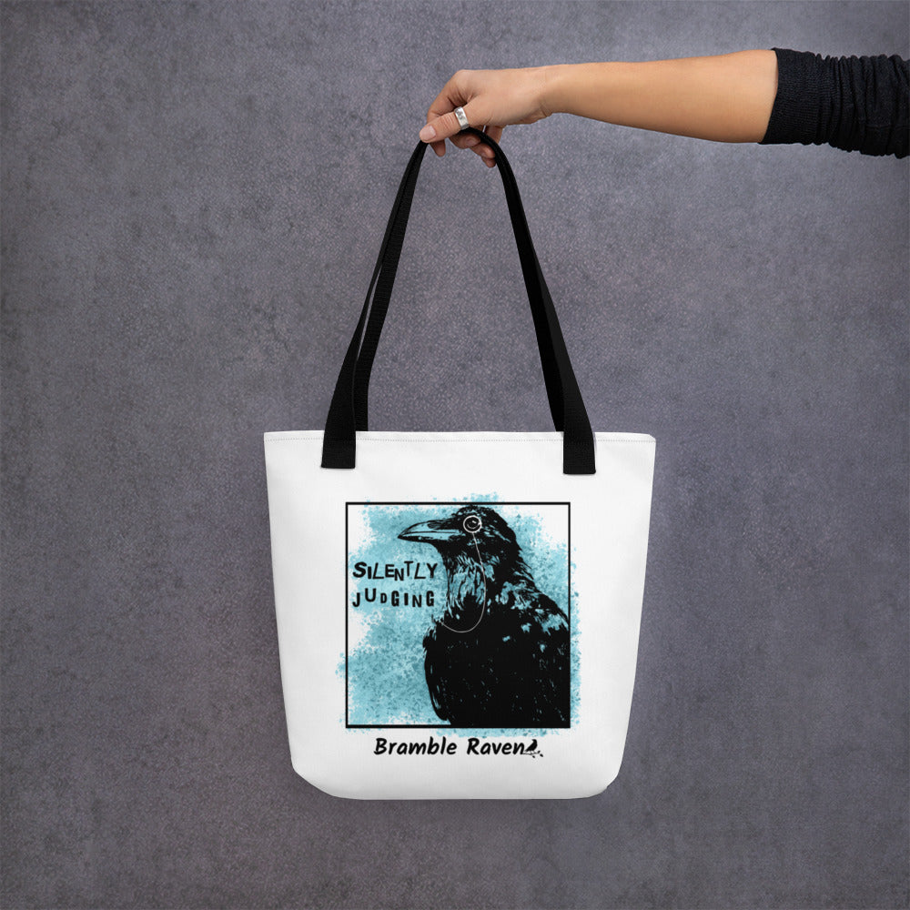15 by 15 inch tote bag with silently judging text by black crow wearing a monocle in a square with blue paint splatters.  Design on both sides of bag. White polyester fabric with black handles. Shown hanging from a model's hand.