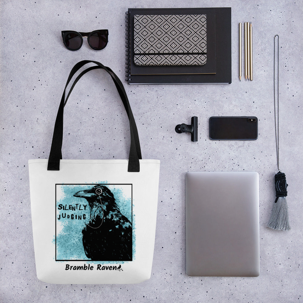 15 by 15 inch tote bag with silently judging text by black crow wearing a monocle in a square with blue paint splatters.  Design on both sides of bag. White polyester fabric with black handles. Shown on tabletop surrounded by items including sunglasses, notebooks, pencils, tablet, phone, and necklace.