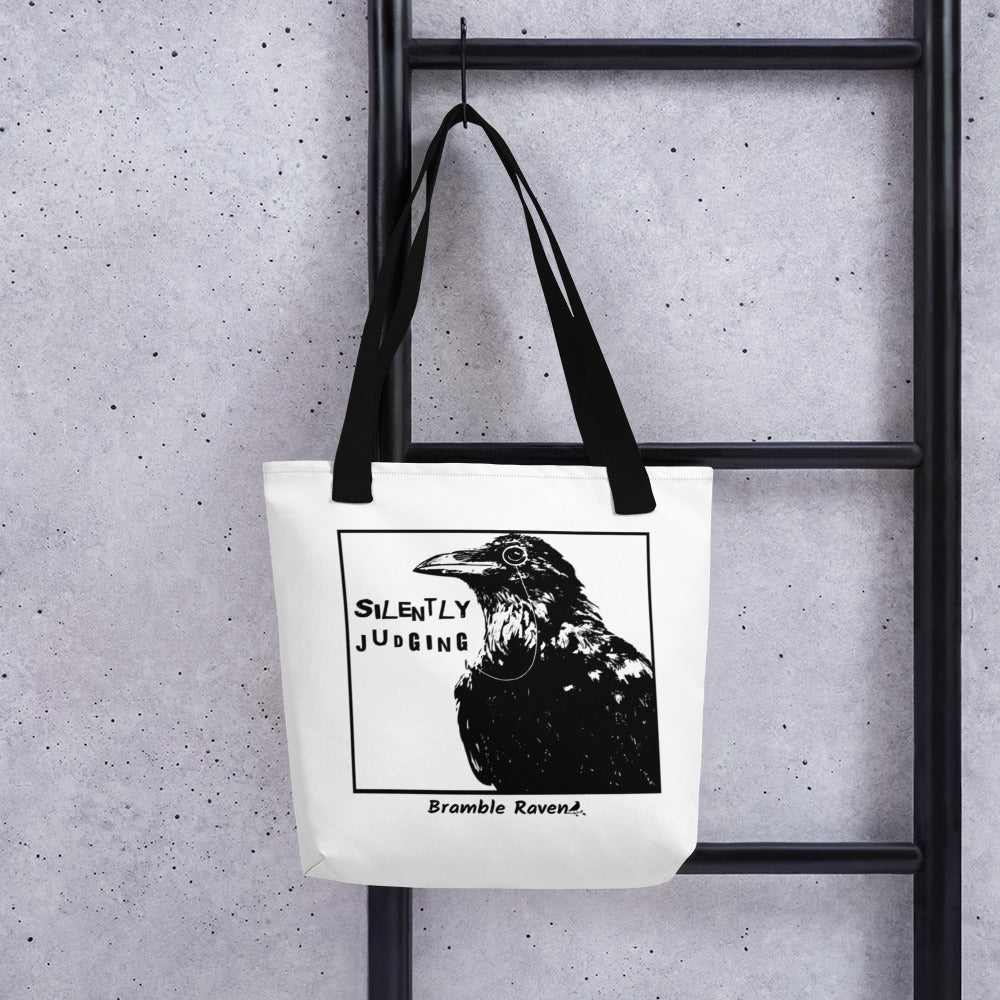 15 by 15 inch polyester tote. Features silently judging text next to black crow wearing a monocle in a square frame on a white background. Image on both sides. white tote bag with black handles. Shown hanging on a black ladder.
