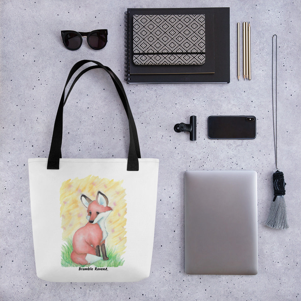 15 by 15 inch white polyester tote bag with black handles. Features original watercolor painting of a fox in the grass against a yellow background. Shown on table surrounded by items including sunglasses, notebooks, pencils, phone, tablet, and necklace.