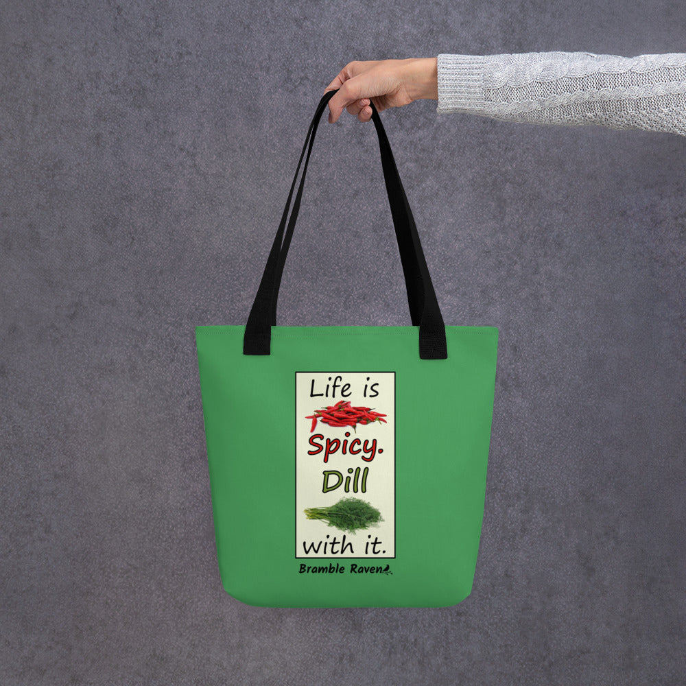 Life is spicy. Dill with it. Phrase with images of chili peppers and dill weed. Rectangular frame for saying on a green background. Polyester tote bag with black handles. 15 by 15 inches. Double sided image. Shown being held by model's hand.