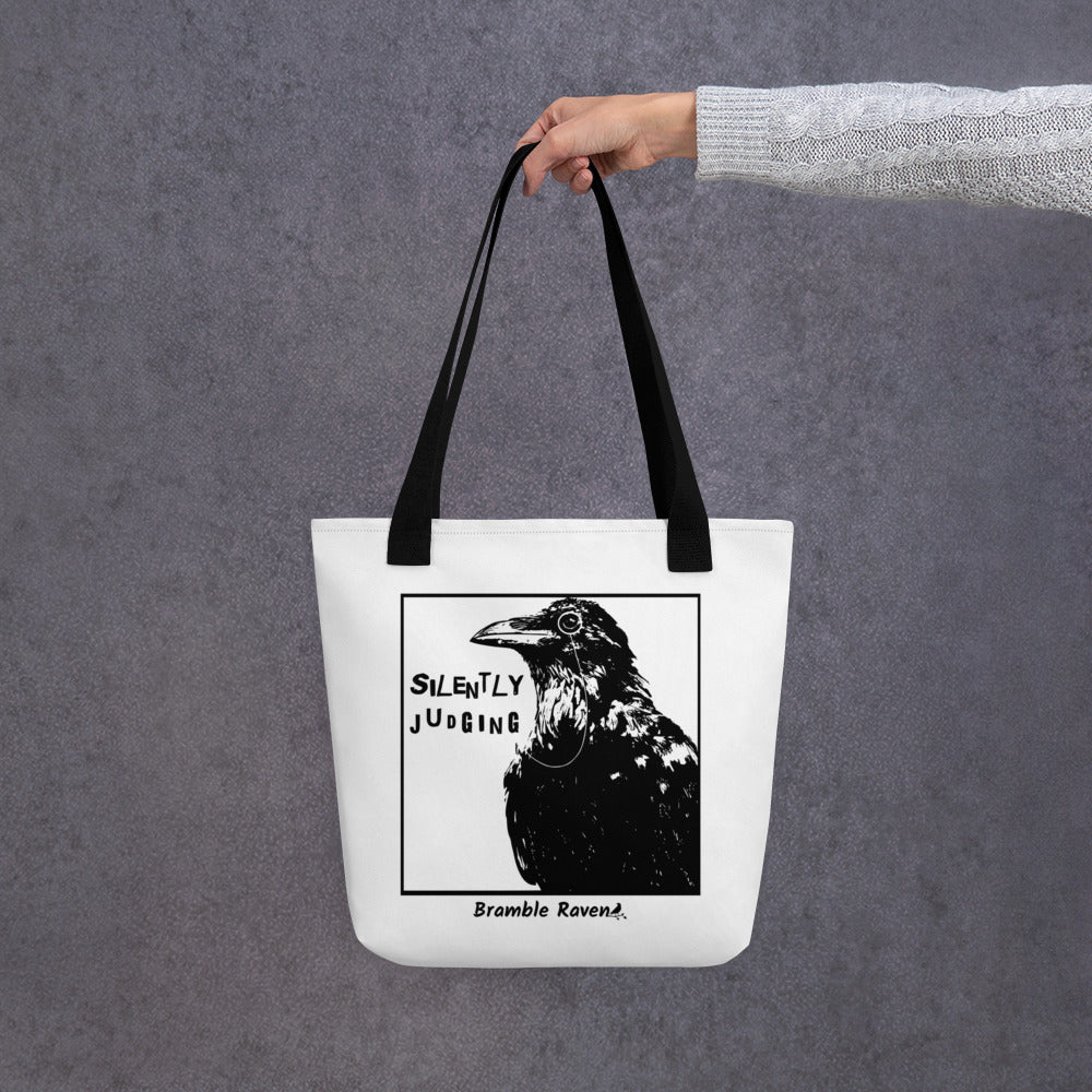 15 by 15 inch polyester tote. Features silently judging text next to black crow wearing a monocle in a square frame on a white background. Image on both sides. white tote bag with black handles. Shown hanging from a model's hand.