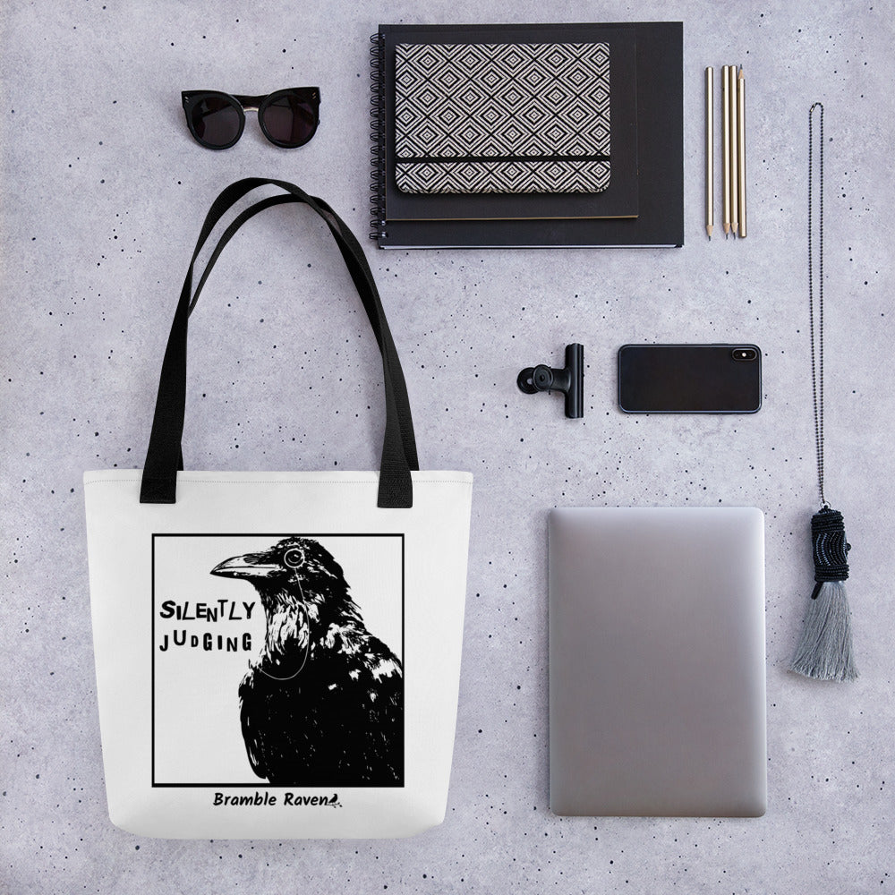 15 by 15 inch polyester tote. Features silently judging text next to black crow wearing a monocle in a square frame on a white background. Image on both sides. white tote bag with black handles. Shown hanging on tabletop surrounded by bag items including sunglasses, notebooks, pencils, phone, tablet, and necklace.