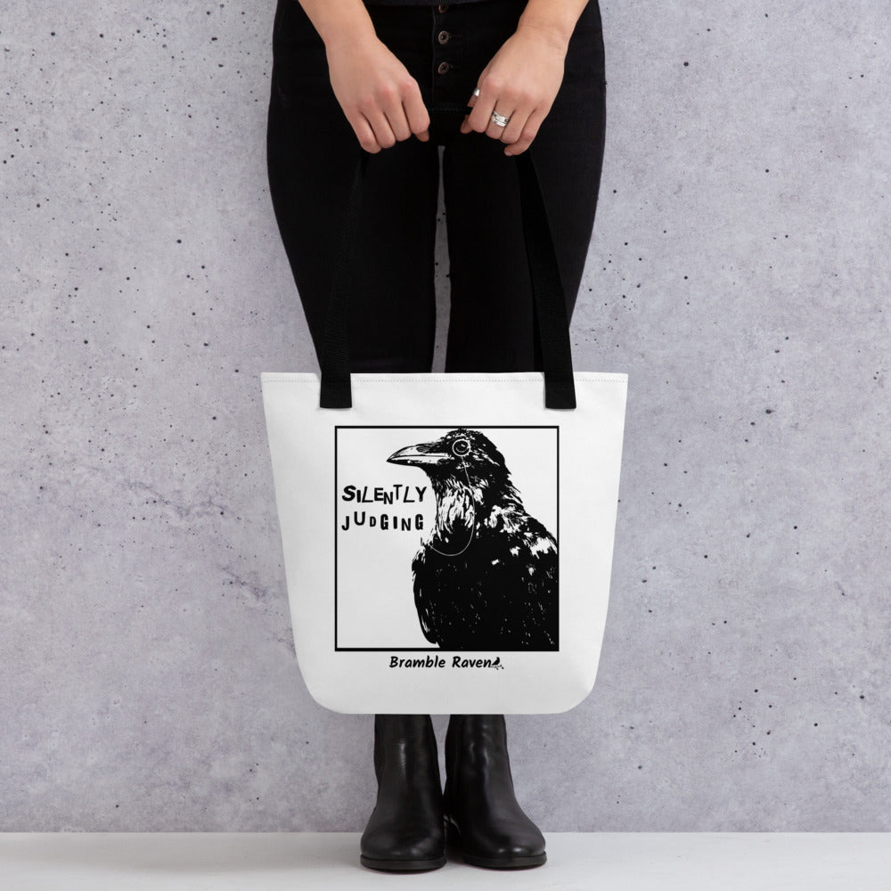 15 by 15 inch polyester tote. Features silently judging text next to black crow wearing a monocle in a square frame on a white background. Image on both sides. white tote bag with black handles. Shown hanging from hands of model.