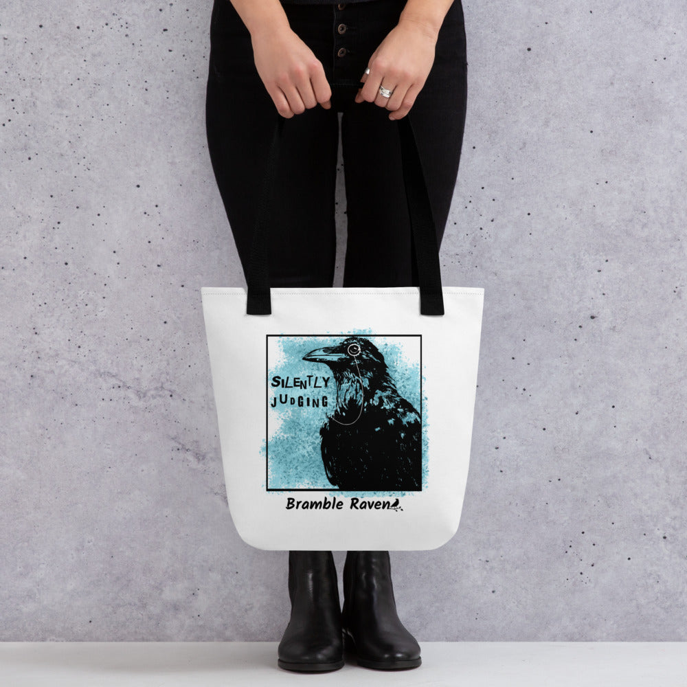 15 by 15 inch tote bag with silently judging text by black crow wearing a monocle in a square with blue paint splatters.  Design on both sides of bag. White polyester fabric with black handles. Shown being held by model.