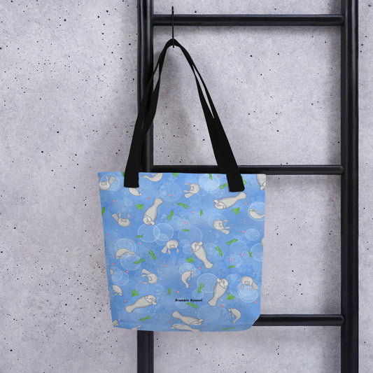 15 by 15 inch polyester tote bag with dual black handles. It has a patterned design of manatees, seashells, seaweed and bubbles on a blue background. Shown hanging from a black ladder.