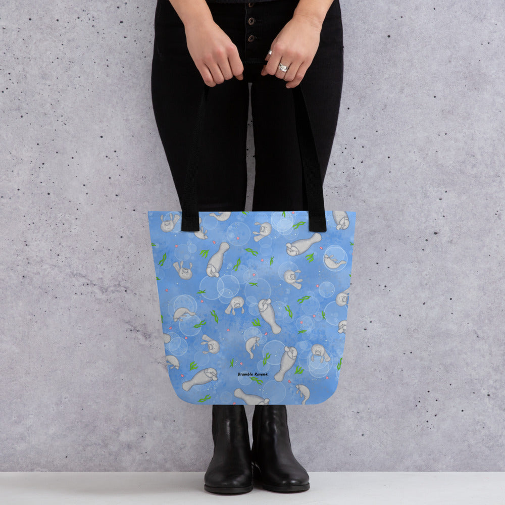 15 by 15 inch polyester tote bag with dual black handles. It has a patterned design of manatees, seashells, seaweed and bubbles on a blue background. Shown hanging from the hands of a model.