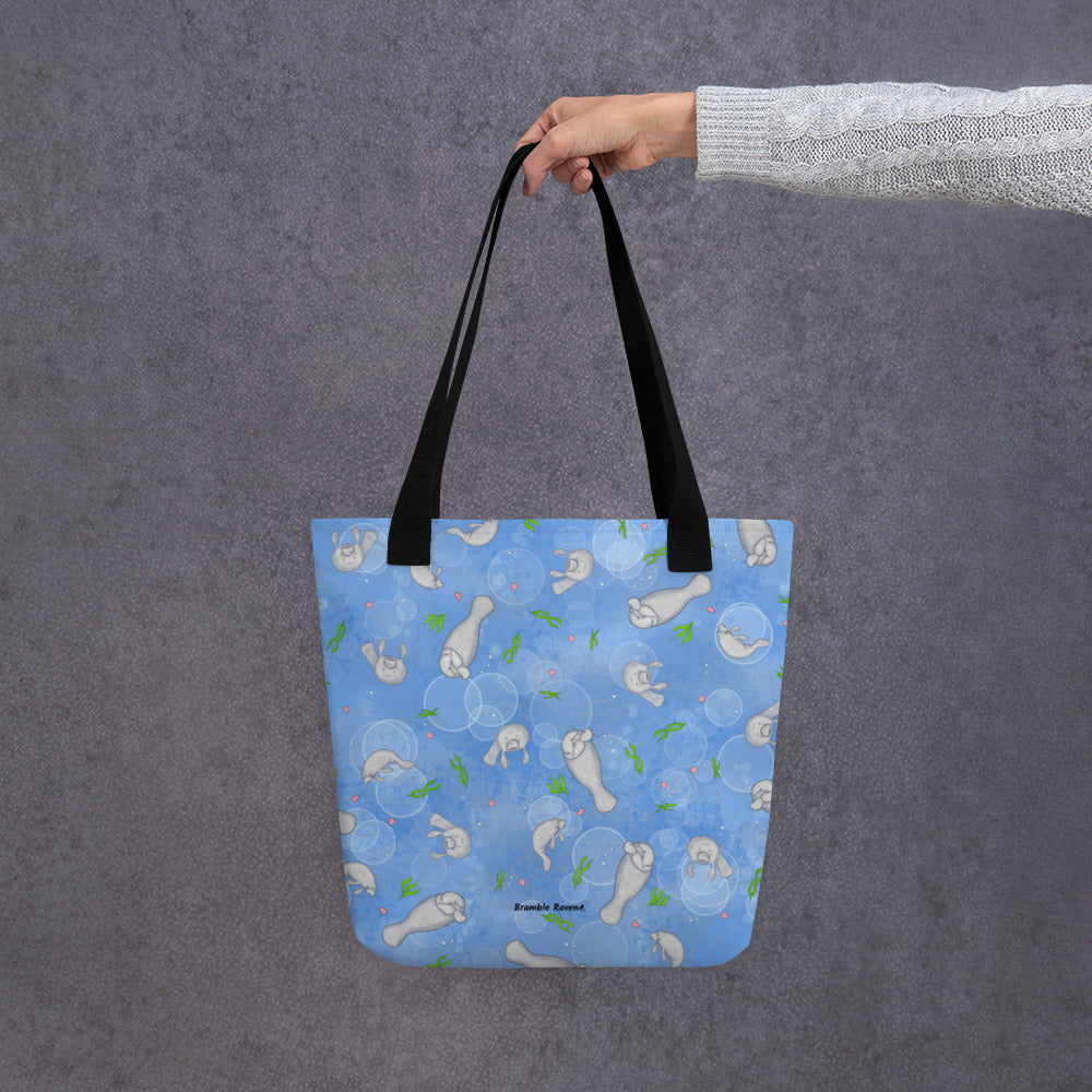 15 by 15 inch polyester tote bag with dual black handles. It has a patterned design of manatees, seashells, seaweed and bubbles on a blue background. Shown hanging from a model's hand.