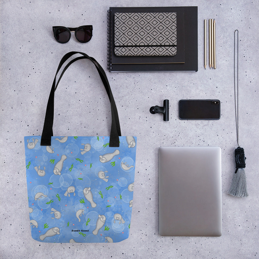 15 by 15 inch polyester tote bag with dual black handles. It has a patterned design of manatees, seashells, seaweed and bubbles on a blue background. Shown on table surrounded by a tablet, notebook, pencils, sunglasses, a phone and a necklace.