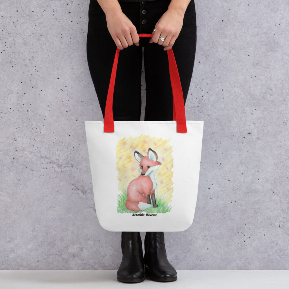 15 by 15 inch white polyester tote bag with red handles. Features original watercolor painting of a fox in the grass against a yellow background. Shown hanging from a model's hands.