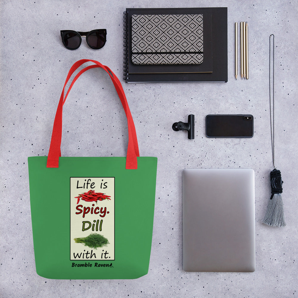 Life is spicy. Dill with it. Phrase with images of chili peppers and dill weed. Rectangular frame for saying on a green background. Polyester tote bag with red handles. 15 by 15 inches. Double sided image. Shown on table surrounded by bag items such as sunglasses, clutch, pencils, phone, tablet, clip and keychain.