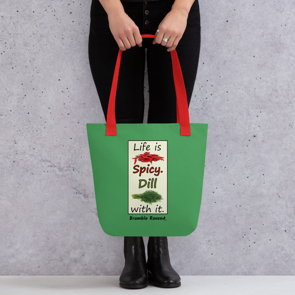 Life is spicy. Dill with it. Phrase with images of chili peppers and dill weed. Rectangular frame for saying on a green background. Polyester tote bag with red handles. 15 by 15 inches. Double sided image. Shown being held by model.