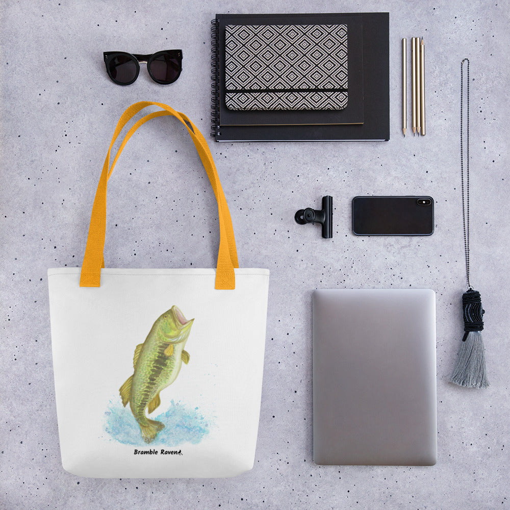 White polyester 15 by 15 inch tote bag. Features a watercolor painting of a largemouth bass leaping out of the water. Yellow handles. Shown hanging from a black ladder. Shown on tabletop surrounded by items including sunglasses, notebooks, pencils, a phone, a tablet, and a necklace.