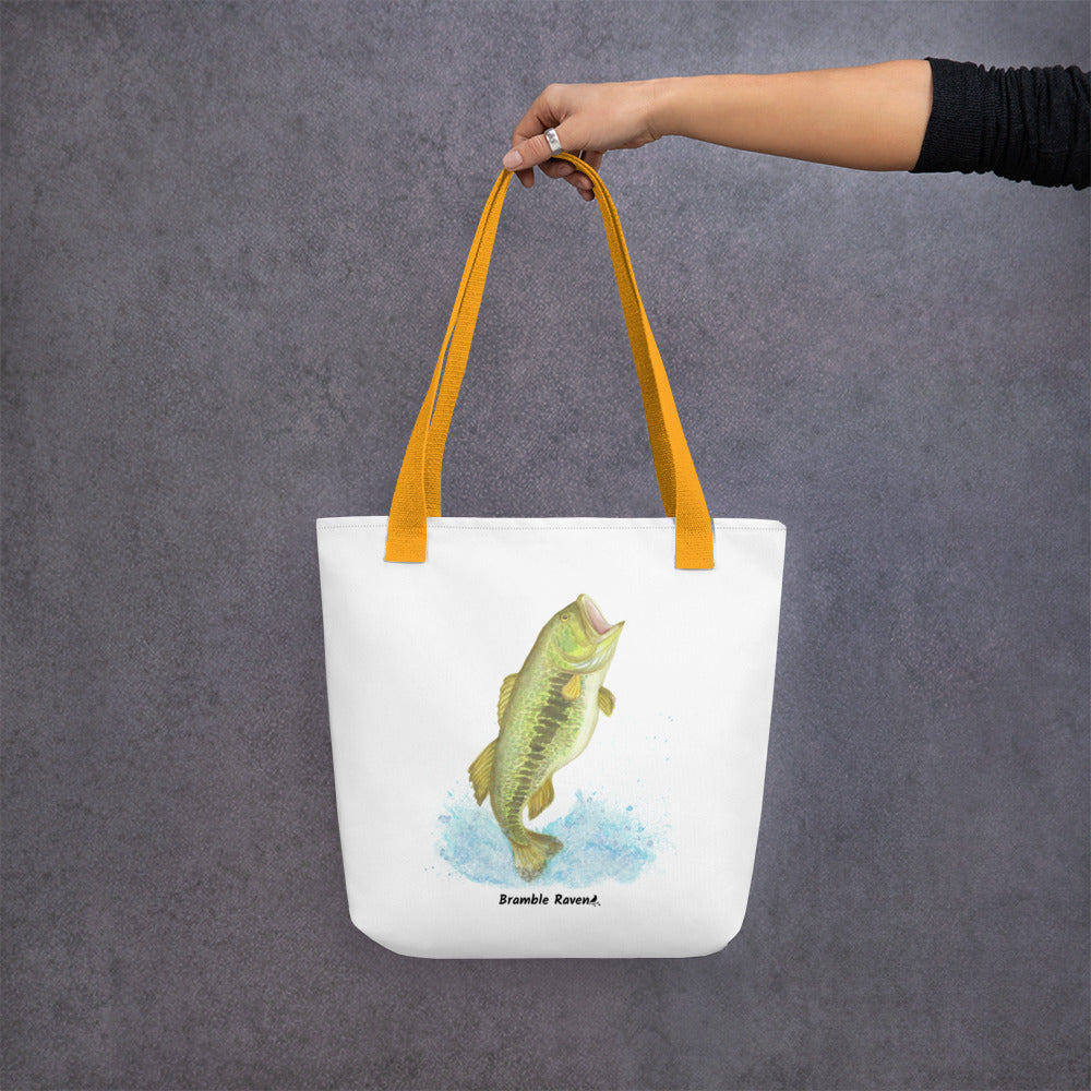 White polyester 15 by 15 inch tote bag. Features a watercolor painting of a largemouth bass leaping out of the water. Yellow handles. Shown hanging from a model's hand.