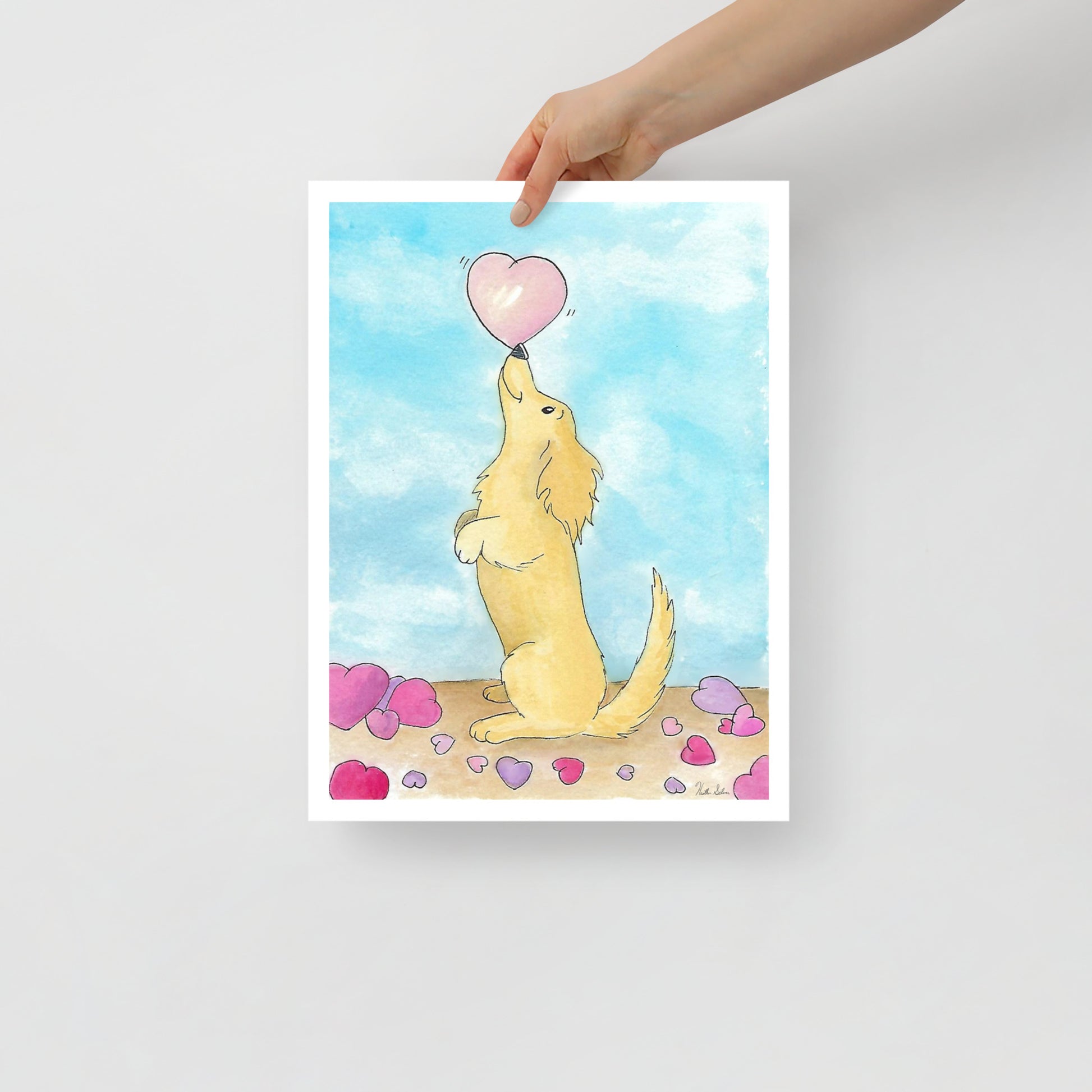 12 x 16 inch enhanced matte paper art print. Watercolor painting entitled Puppy love. Design shows a cute puppy balancing a heart on its nose, with hearts on the floor and a blue sky background. Shown being held by model's hand.