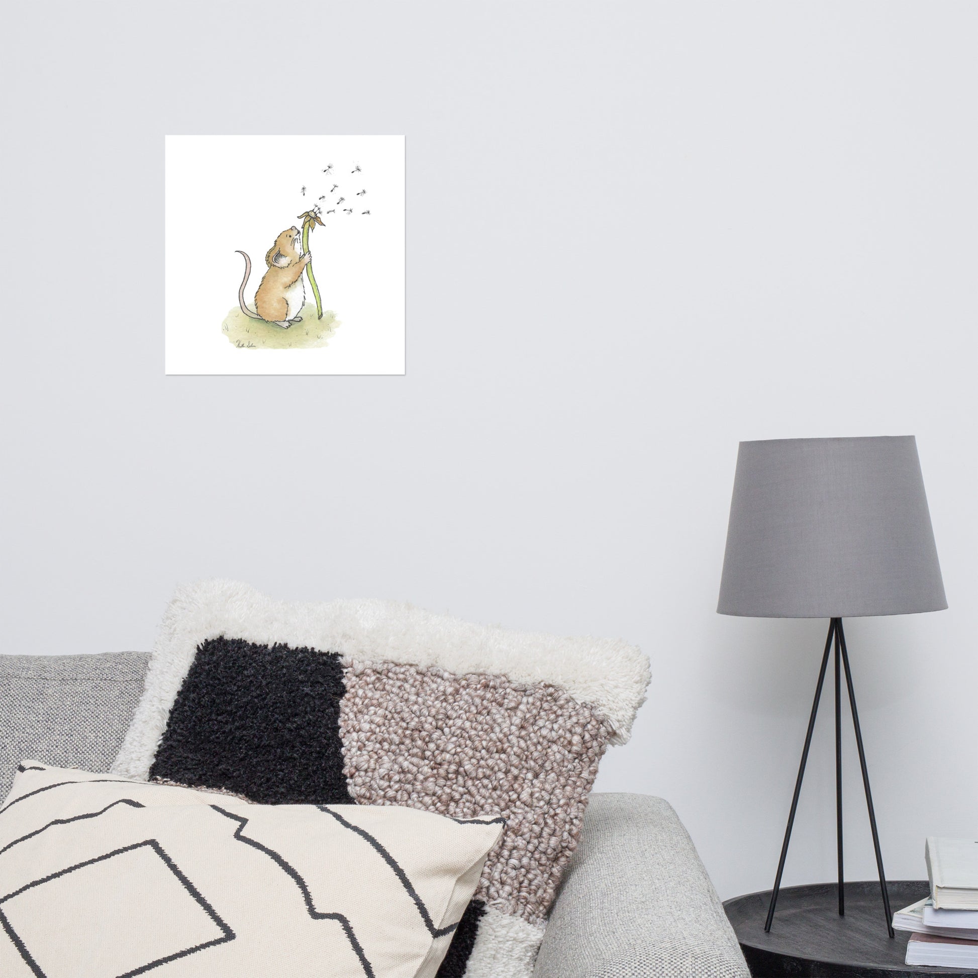 14x14 matte paper art print. Giclée quality printing. Design is a watercolor painting of a cute mouse blowing the seeds off a dandelion stem. Shown on the wall above a grey couch and lamp.