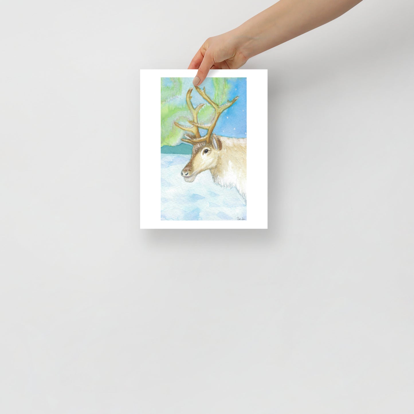 11 by 14 inch art poster print. Watercolor painting of a snowy reindeer with the northern lights in the background. Shown being held by a model's hand.