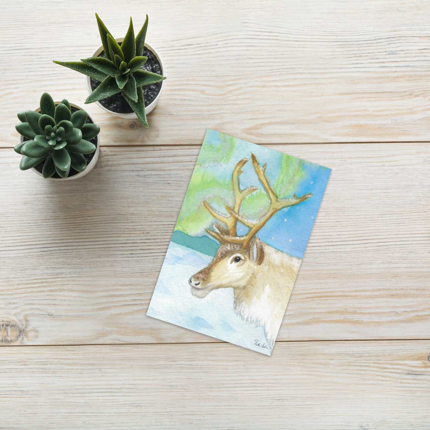4 x 6 inch greeting card. Features watercolor print of a reindeer in the snow with northern lights in the sky. Blank inside. Comes with a white envelope. Shown on table by potted plants.