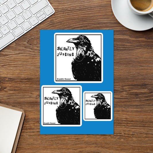 Glossy sticker sheet with three different sizes of kiss cut stickers with white borders on a single sheet. Features the silently judging text by black crow wearing a monocle in a square on a white background. Shown on tabletop.