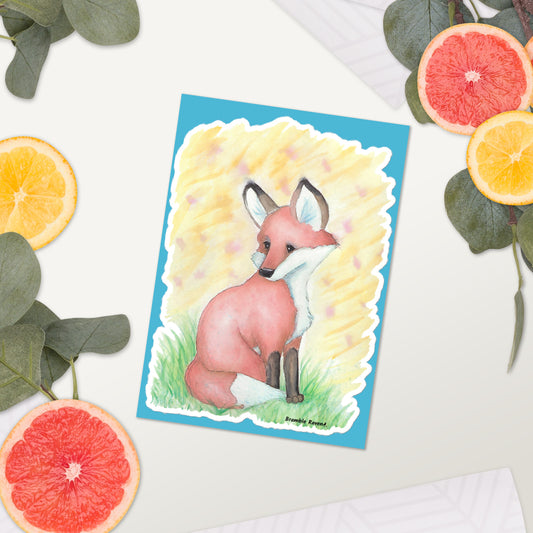 Glossy kiss-cut sticker with white border. Features original watercolor painting of a fox in the grass against a yellow background. Shown on tabletop surrounded by citrus slices and leaves.