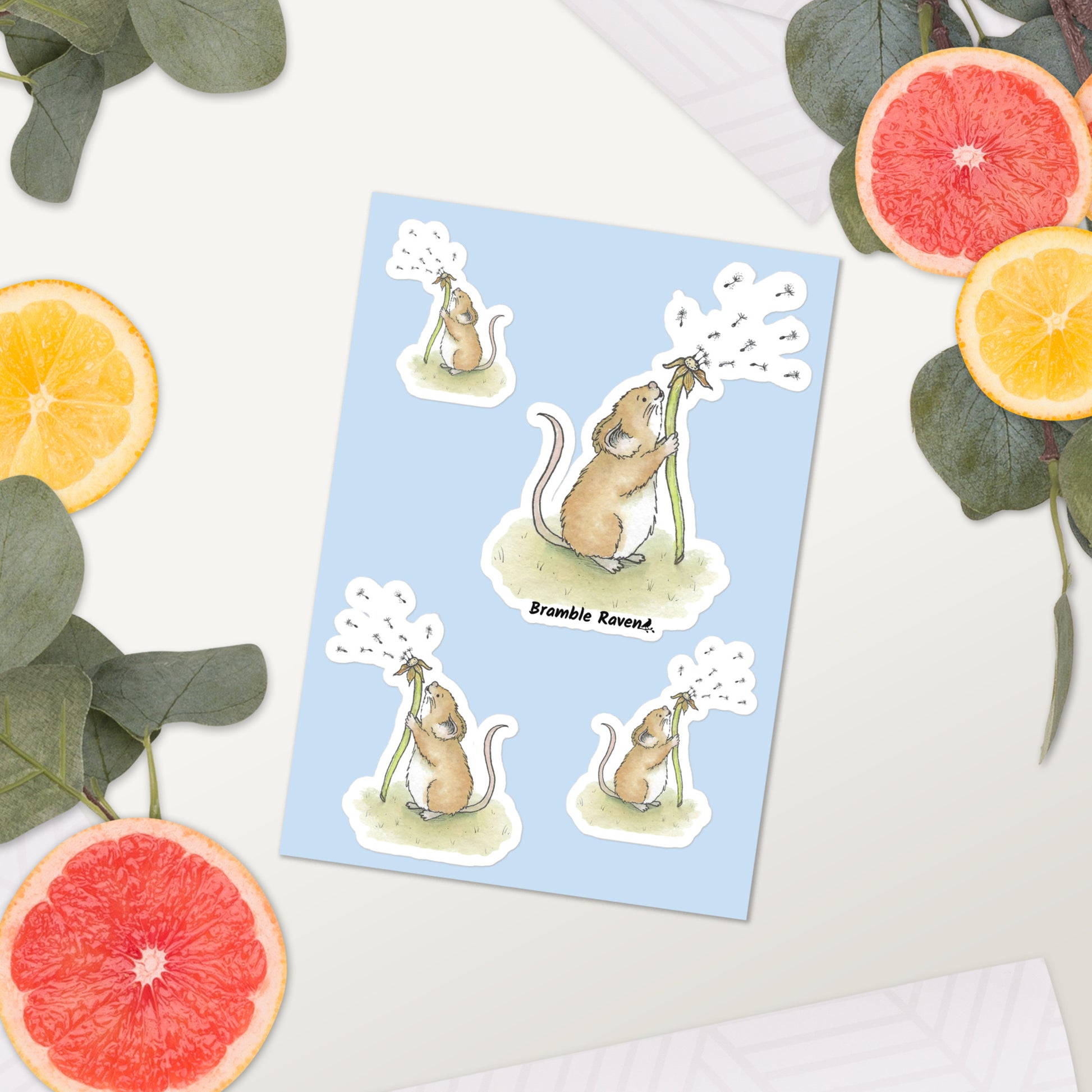 Original Dandelion Wish design of cute watercolor mouse blowing dandelion seeds. Glossy sticker sheet with four mice in different sizes and directions. Each sticker is kiss cut with a white border.  Shown on table surrounded by citrus slices.