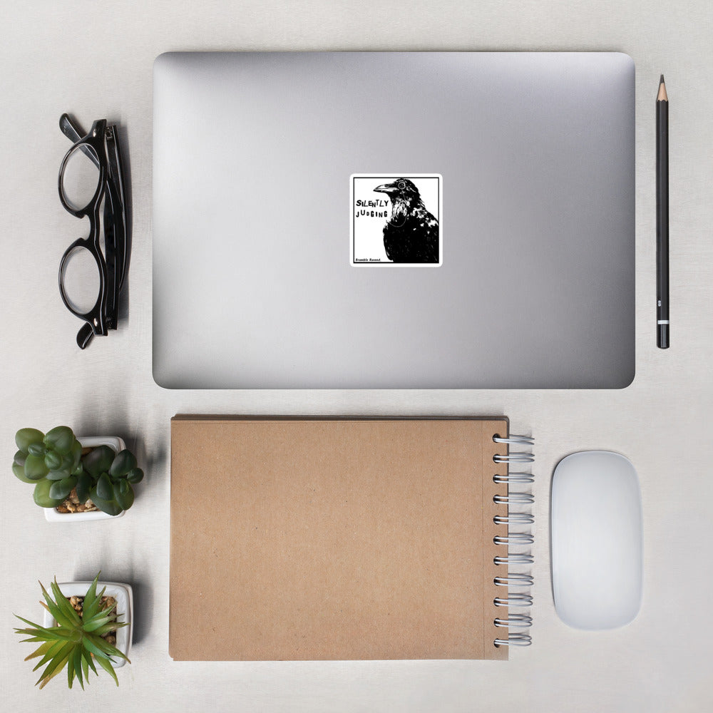 3 inch kiss-cut vinyl sticker with white border.. Features silently judging text next to black crow wearing a monocle in a square frame on a white background.  Shown on laptop.