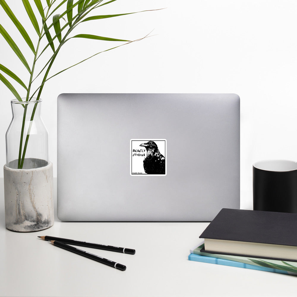 3 inch kiss-cut vinyl sticker with white border.. Features silently judging text next to black crow wearing a monocle in a square frame on a white background.  Shown on laptop.