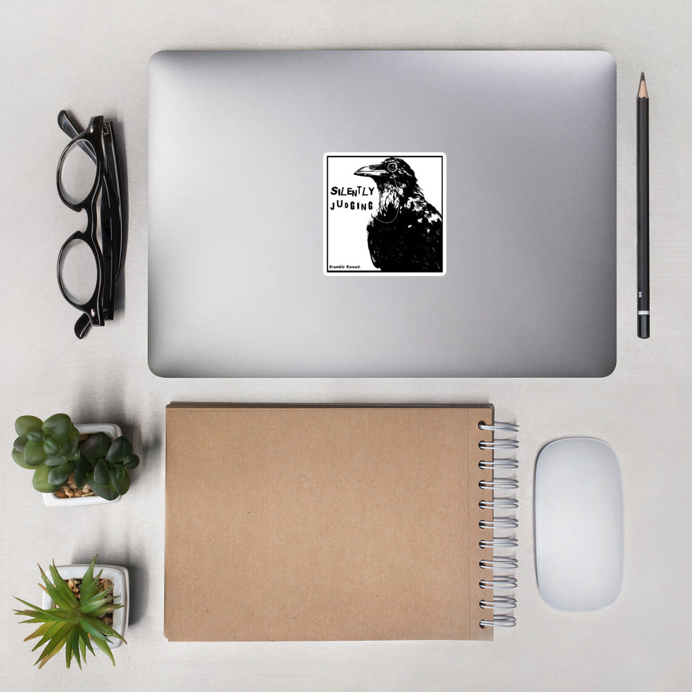 4 inch kiss-cut vinyl sticker with white border.. Features silently judging text next to black crow wearing a monocle in a square frame on a white background.  Shown on laptop.