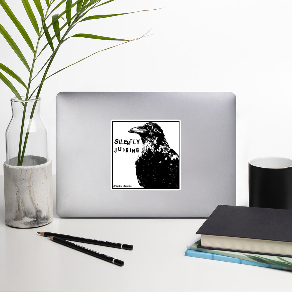 5.5 inch kiss-cut vinyl sticker with white border.. Features silently judging text next to black crow wearing a monocle in a square frame on a white background.  Shown on laptop.