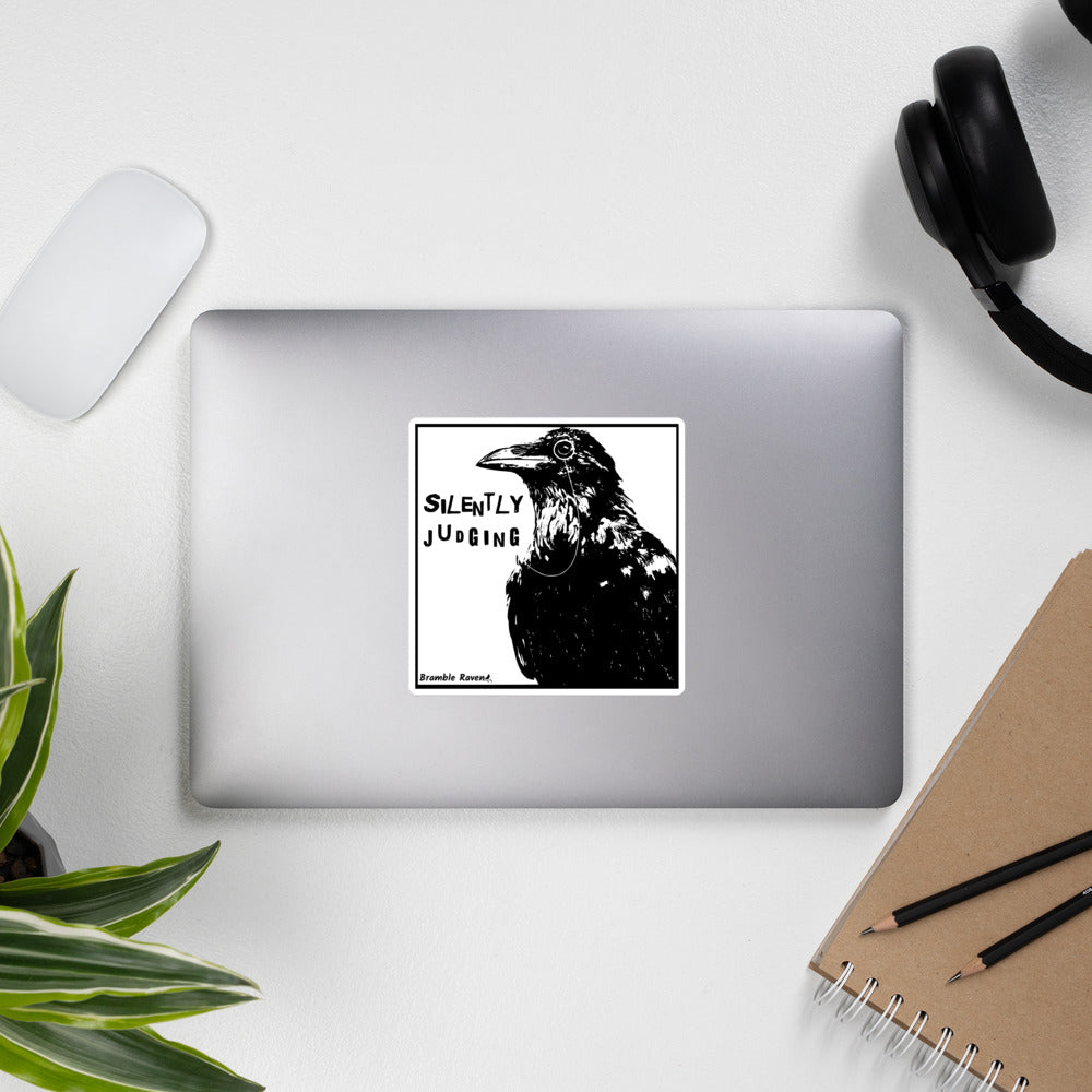 5.5 inch kiss-cut vinyl sticker with white border.. Features silently judging text next to black crow wearing a monocle in a square frame on a white background.  Shown on laptop.