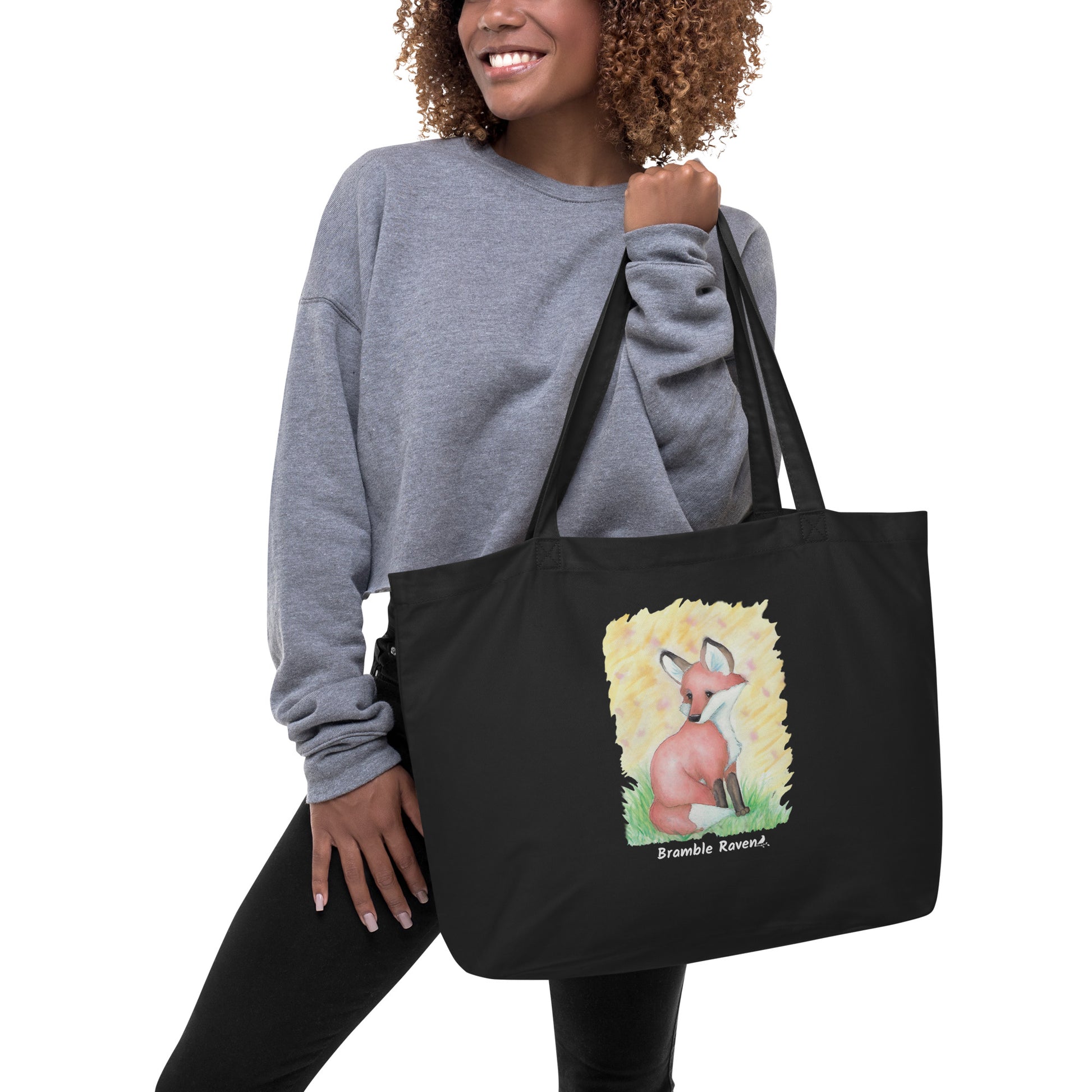 Original watercolor fox painting in the grass with yellow background. Printed on large black colored eco tote. 20 by 14 by 5 inches. 100% recycled cotton. Shown being held by female model.