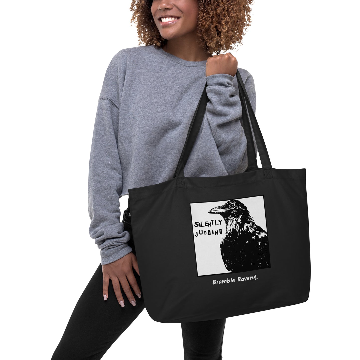 Original Silently Judging crow design. Silently Judging text. Black crow with monocle in square frame. Printed on large black colored eco tote. 20 by 14 by 5 inches. 100% recycled cotton. Shown being held by female model.