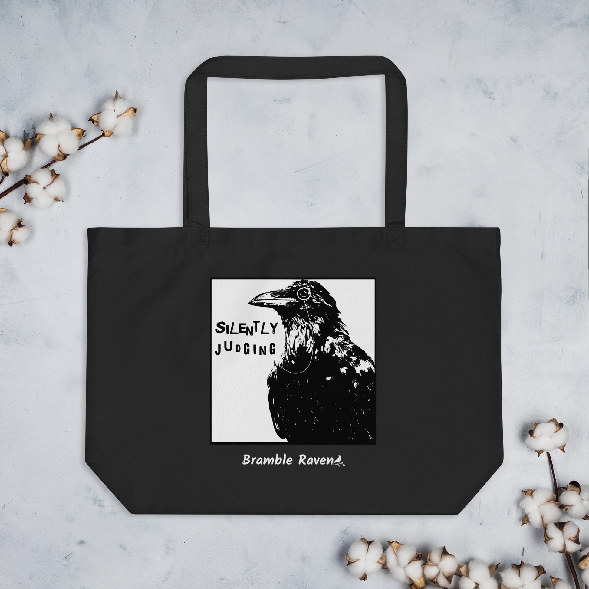 Original Silently Judging crow design. Silently Judging text. Black crow with monocle in square frame. Printed on large black colored eco tote. 20 by 14 by 5 inches. 100% recycled cotton. Shown on tabletop with cotton plant accents.