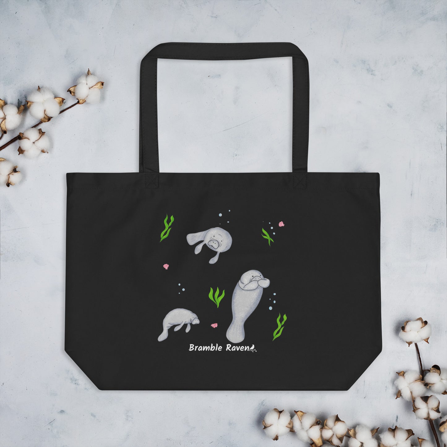 20″ × 14″ × 5″ 100% organic cotton tote bag. Features three manatees with accents of seashells, seaweed, and bubbles. Black fabric with dual handles. Shown on table with sprigs of cotton.